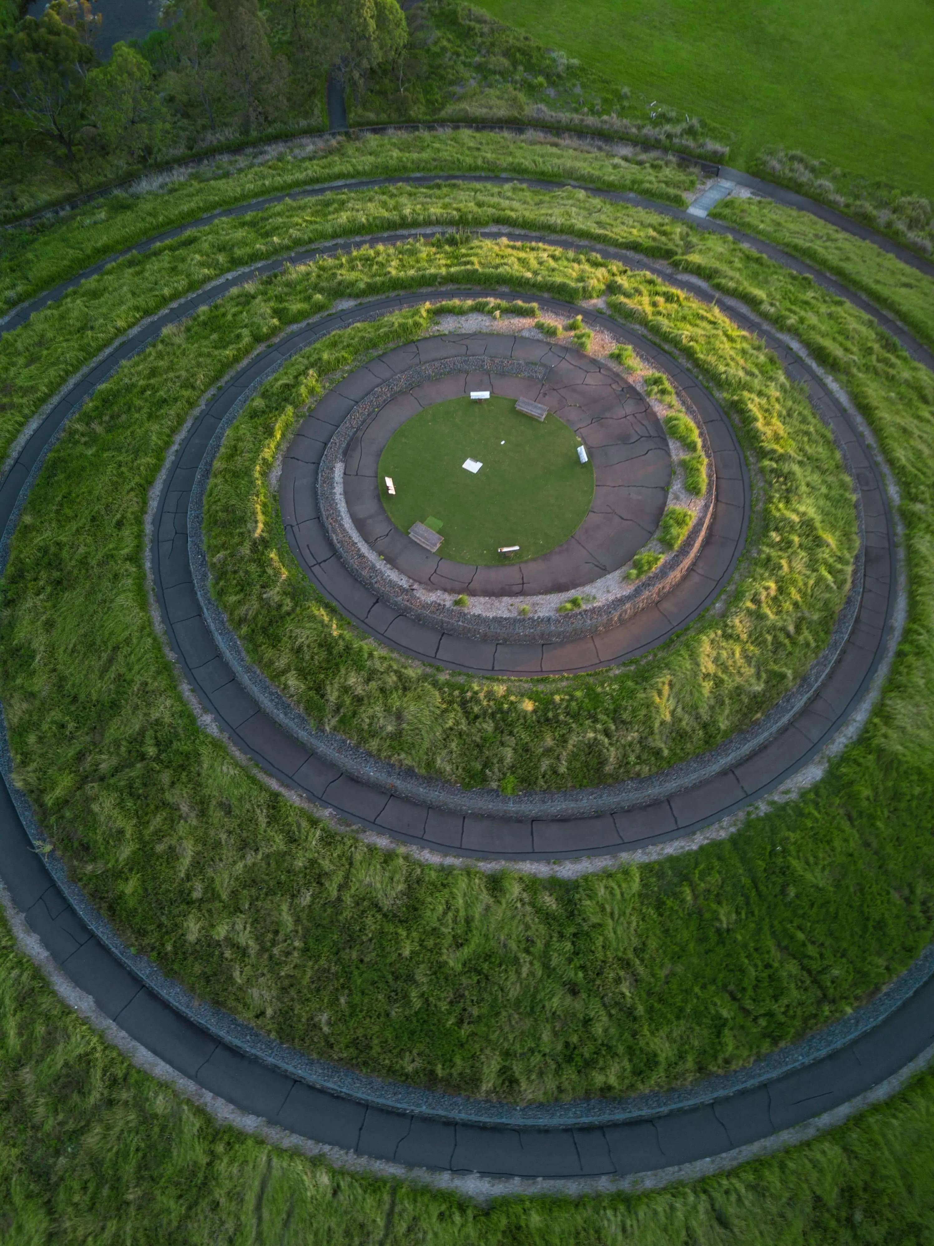 Aerial view of a circular, spiral-shaped landscape with green grass and paved walkways leading toward a central seating area. The design resembles a labyrinth or meditation garden, creating a peaceful and harmonious natural setting.